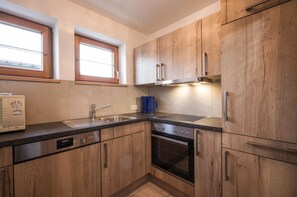 A compact kitchen with wooden cabinets, a built-in oven, a dishwasher, and a window above the sink. - Haus Adler 4 by Alpine Host Helpers (Westendorf)