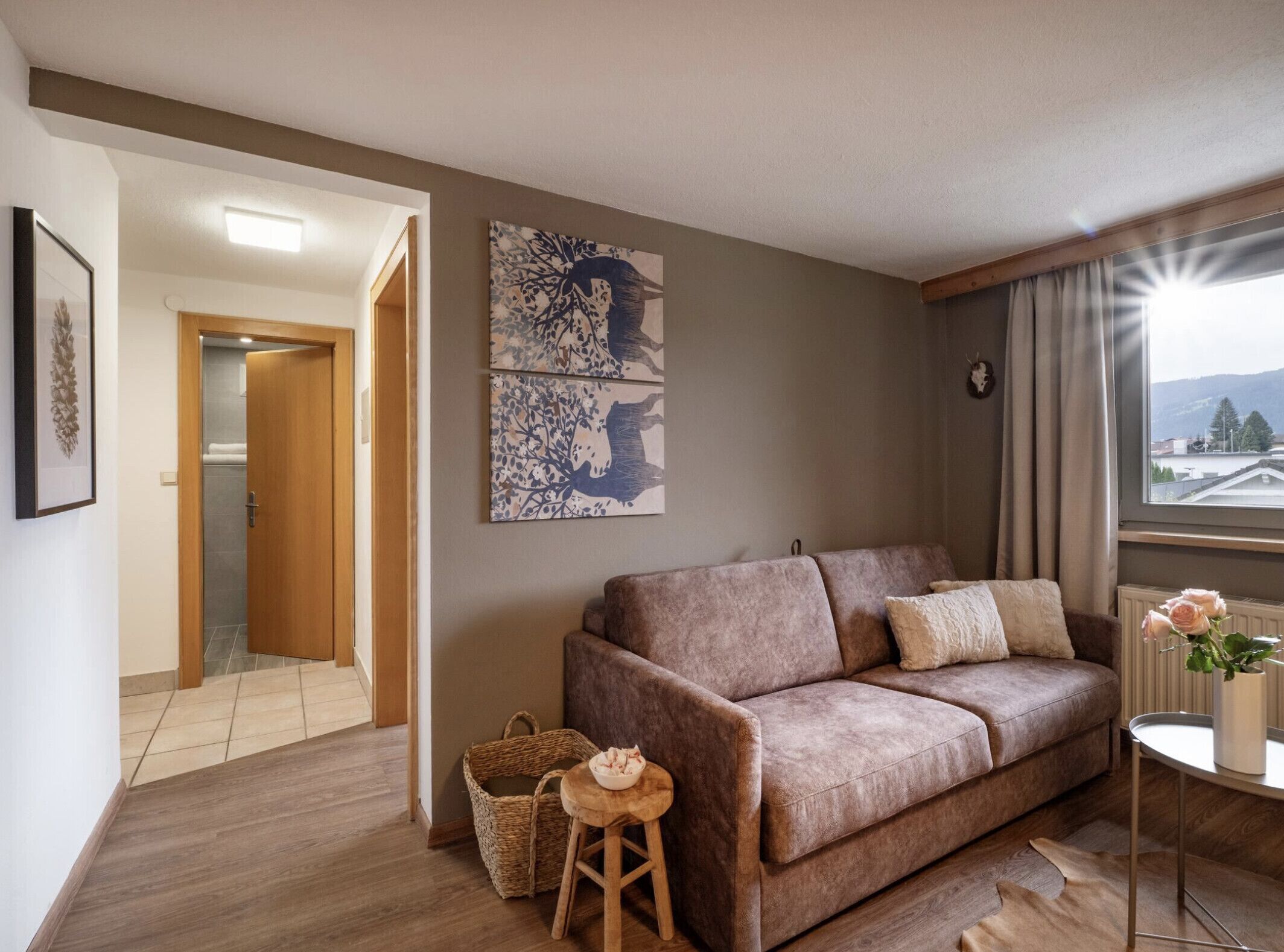 A living room with a brown sofa, a small table with a vase of flowers, and a window with a view of a mountain.