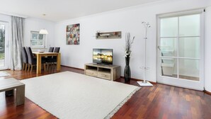 A modern living room with a wooden floor, a dining area with a table and chairs, a flat-screen TV on a wooden stand, a large window with a view, and a coat rack. - Kitz-Heart Standard Top 1 (Kitzbühel)