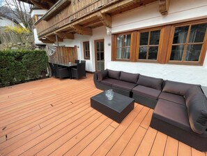 Um deck de madeira com sofá e mesa, cerca de madeira e um edifício com grandes janelas.