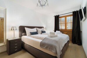 Quarto com cama, mesas de cabeceira, luminária, janela com cortinas e lustre.