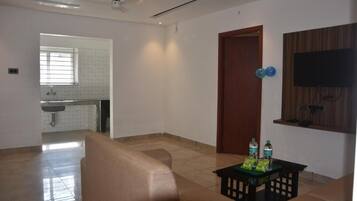 A spacious living area with a sofa, a small table with water bottles, a wooden door, and a TV mounted on the wall.