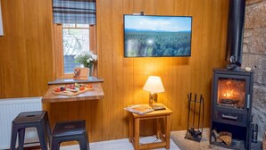 Una habitación acogedora con paredes de madera, un televisor sobre una ventana, una mesa con una bandeja de frutas, una lámpara y un estufa de leña.