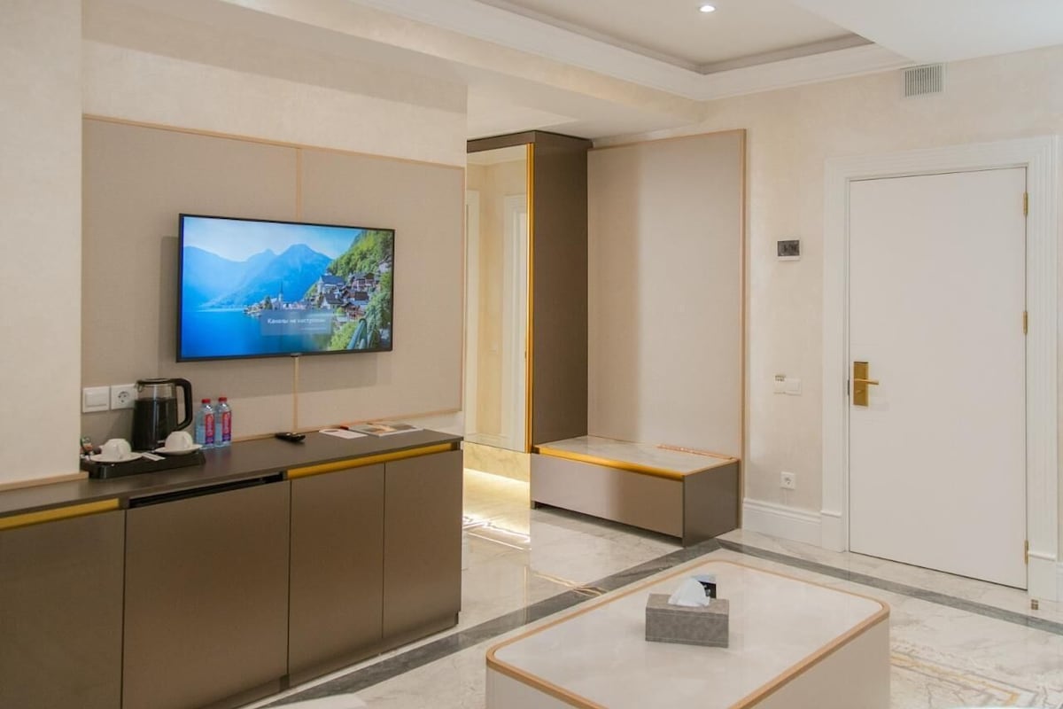 Un vestíbulo de hotel con una recepción, un televisor mostrando una vista panorámica, una puerta blanca y un piso de mármol.