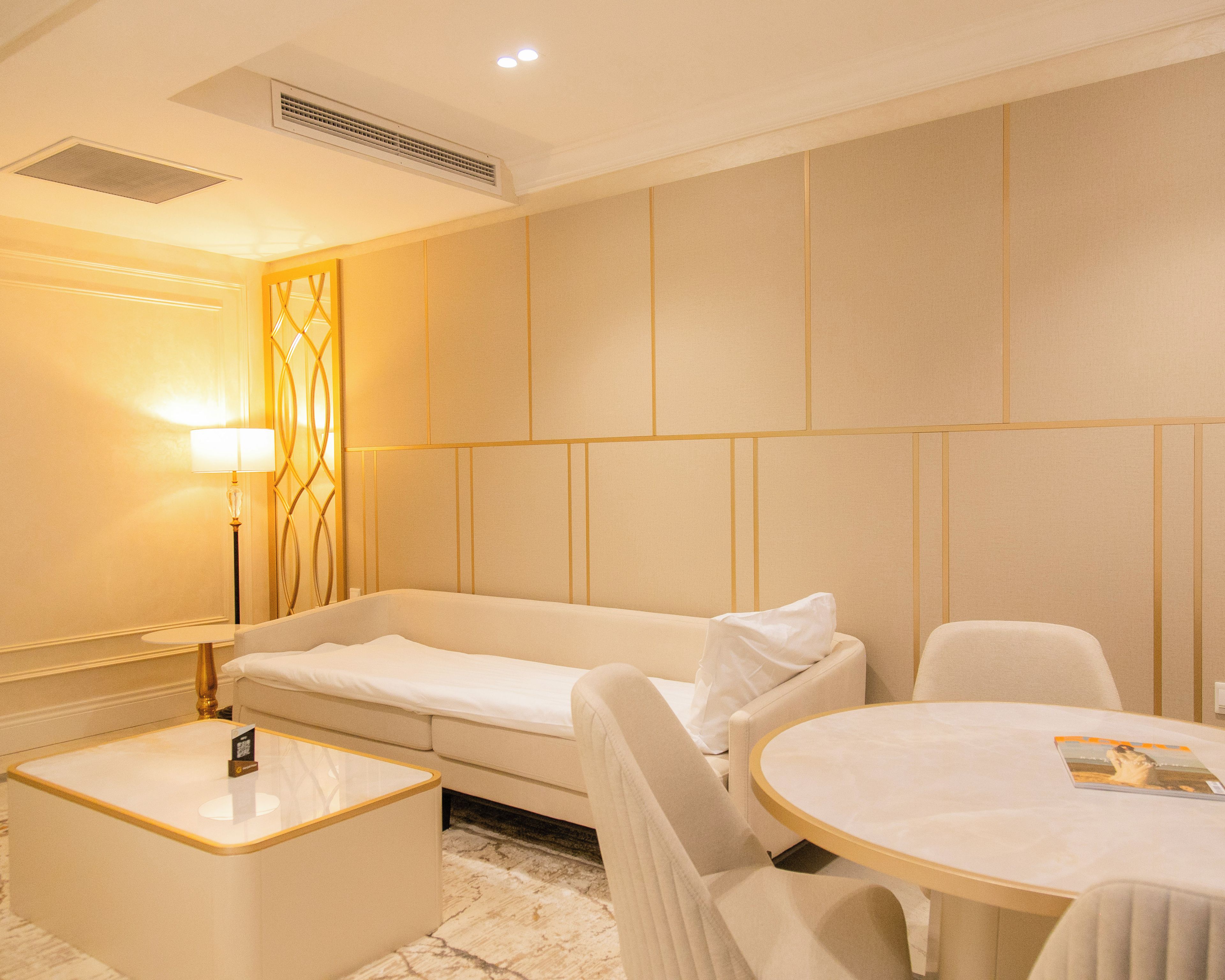 A hotel room with a sofa, a round table, a lamp, and a small bedside table.