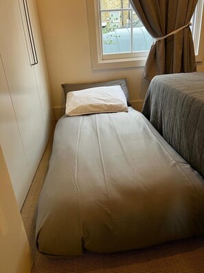 A single bed with a grey bedspread and a white pillow in a room with a window and curtains. - Modern Angel Flat - King Bed & Sofa Bed (London)