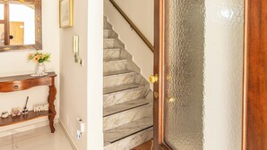 Uma escadaria com degraus de mármore que levam a um patamar, uma porta de madeira com painel de vidro jateado, um console com um vaso e um telefone fixado na parede.