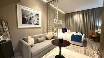 Habitación de hotel con sofá, una mesa redonda, una cama con manta azul y un cuadro grande en la pared.