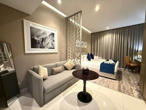 Ein Hotelzimmer mit einer Couch, einem runden Tisch, einem Bett mit blauer Decke und einem großen gerahmten Bild an der Wand.