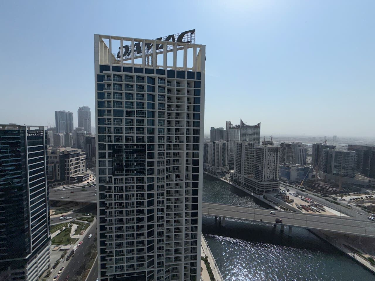 A high-rise building with a grid-like facade, surrounded by other high-rise buildings and a river with a bridge.