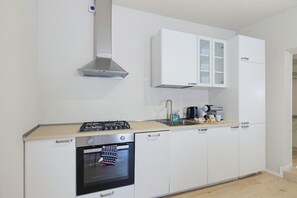 Ett modernt kök med vita skåp, en rostfri köksfläkt, en inbyggd ugn och en bänkskiva med en kaffebryggare och andra köksredskap.