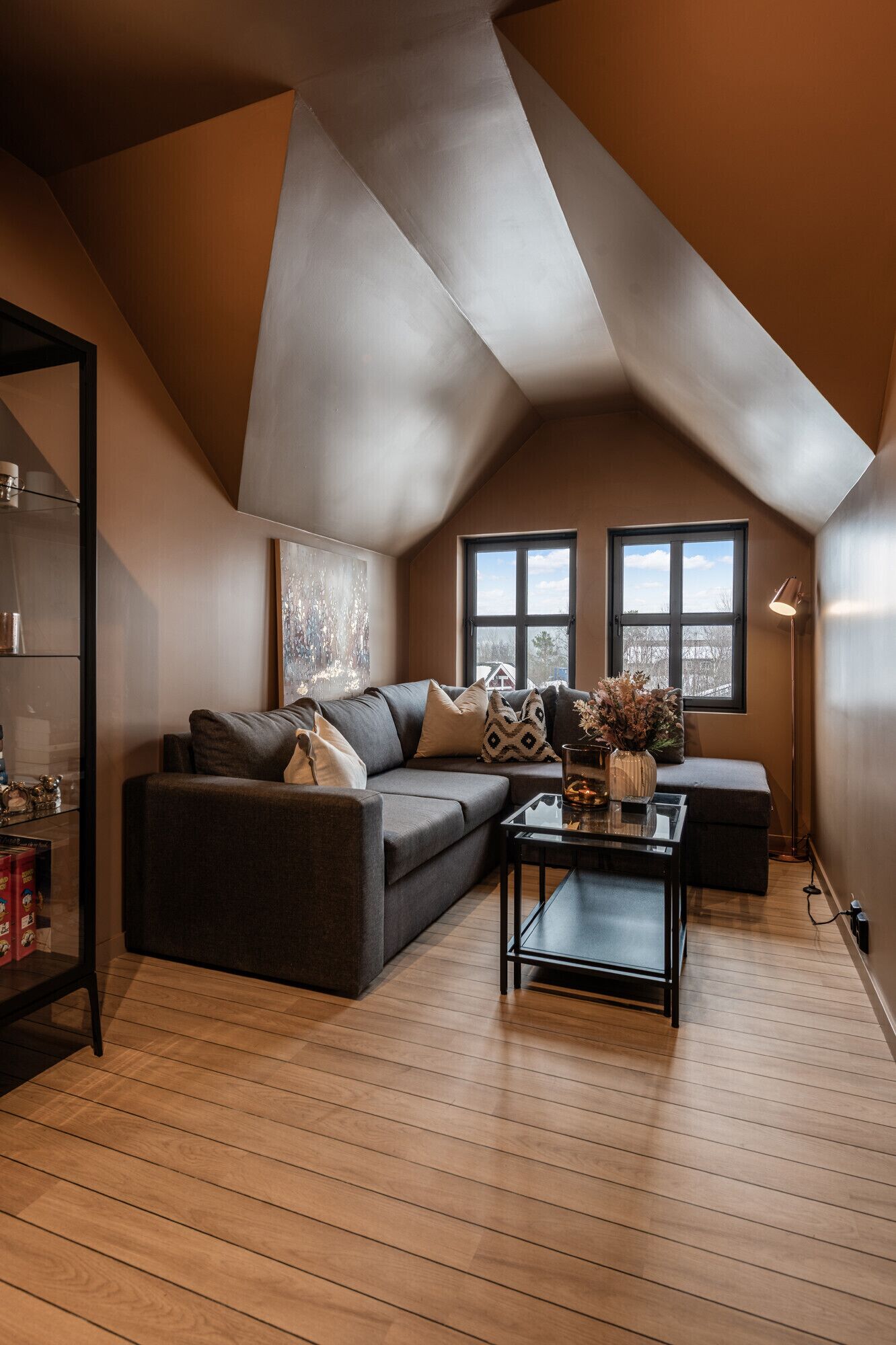 A loft-style living room with a sloped ceiling, a gray sofa, a coffee table, and two windows.