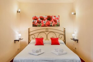 Habitación de hotel con cama, dos almohadas rojas, dos lámparas de noche, un cuadro de flores rojas en la pared y dos toallas dobladas sobre la cama.
