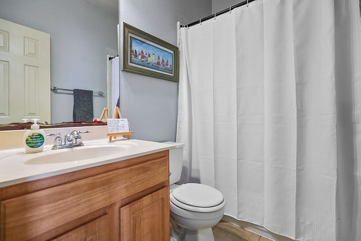 Ein Badezimmer mit einem weißen Waschbecken, einem Holzkabinent, einem gerahmten Bild über dem Spiegel, einer weißen Toilette und einem weißen Duschvorhang.