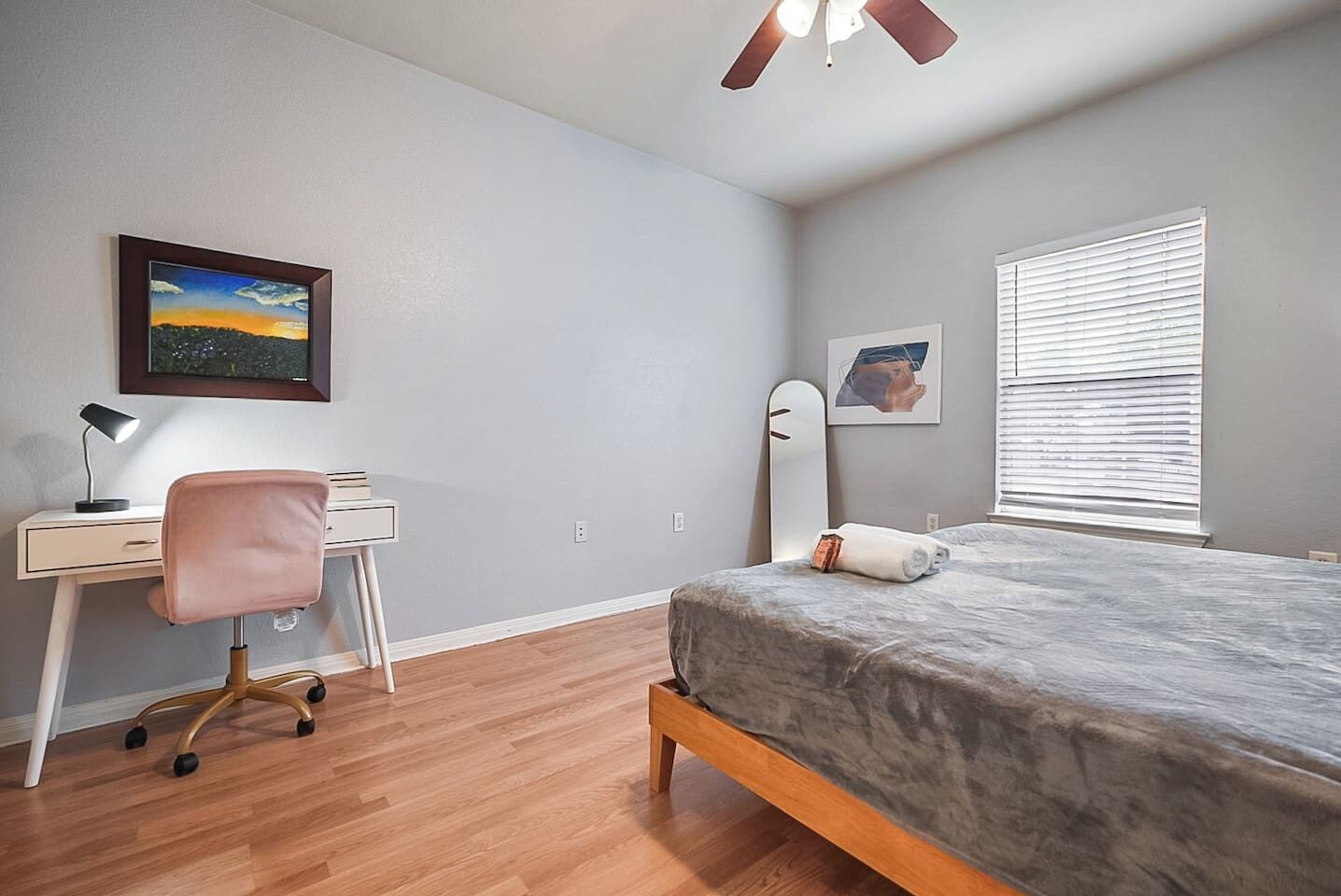 Ein Schlafzimmer mit Bett, Schreibtisch, Stuhl, Deckenventilator, Fenster mit Jalousien und zwei gerahmten Bildern an der Wand.