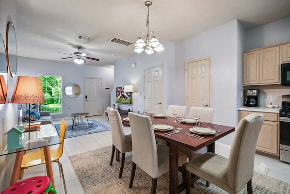 A dining area with a glass table, chairs, and a chandelier. There is a kitchen area with cabinets and appliances.
