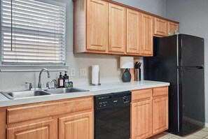 Eine Küche mit Holzschränken, einem schwarzen Kühlschrank, einem Geschirrspüler, einer Spüle mit Wasserhähnen und einem Fenster mit Jalousien.