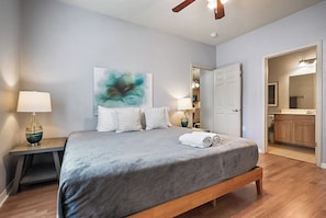 A bedroom with a bed, bedside lamps, a painting, a door to a bathroom, and a wooden floor.