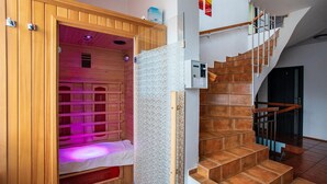 Uma sauna de madeira com um banco e divisória de vidro, adjacente a uma escada com degraus de madeira e corrimão de metal.