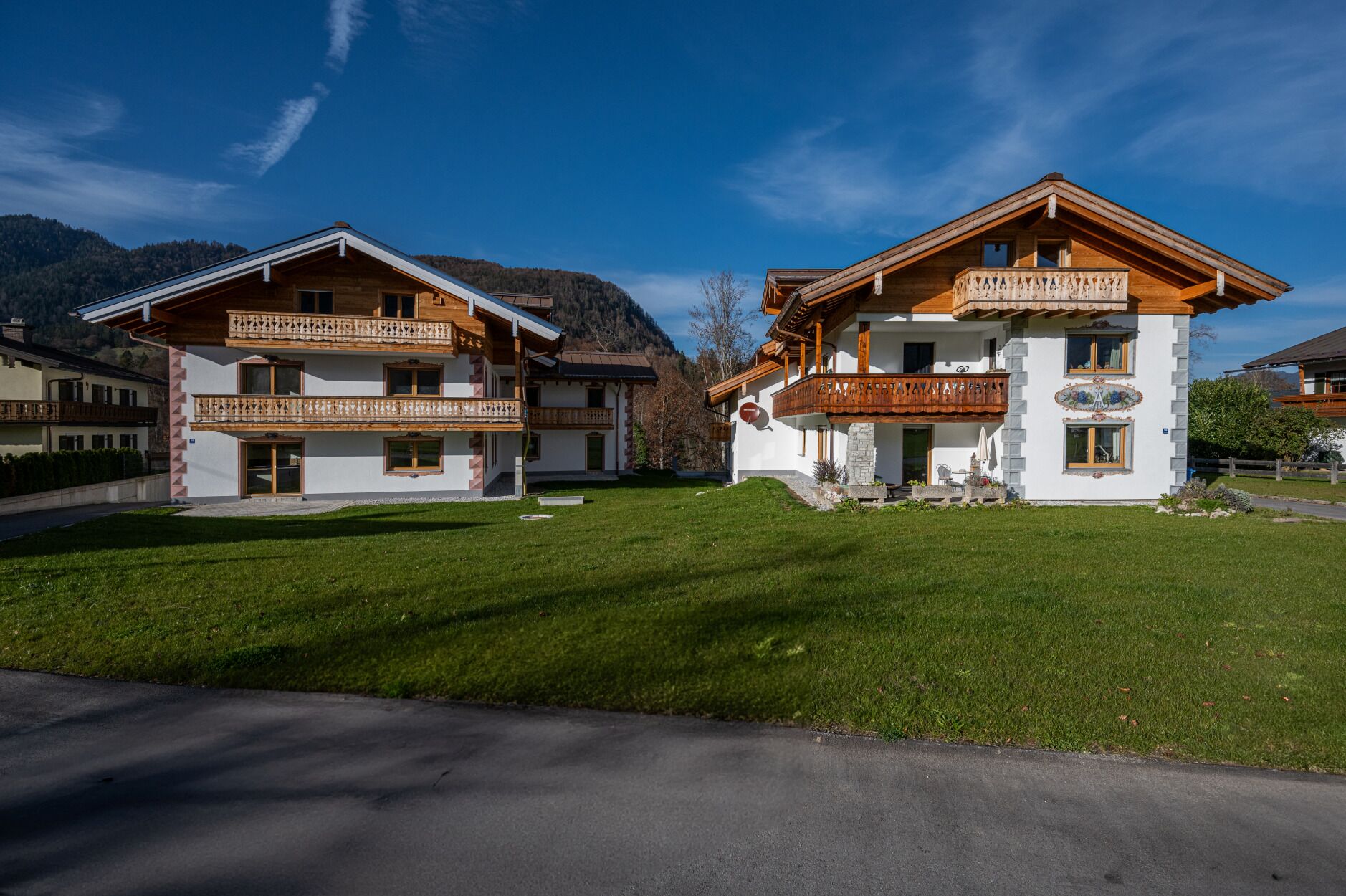 A two-story chalet with a balcony, surrounded by a well-maintained lawn and a mountain backdrop.