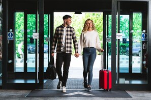 A couple walking through an automatic door with a red suitcase, a black bag, and a mat on the floor. - The Village Hotel Sheffield (Sheffield)