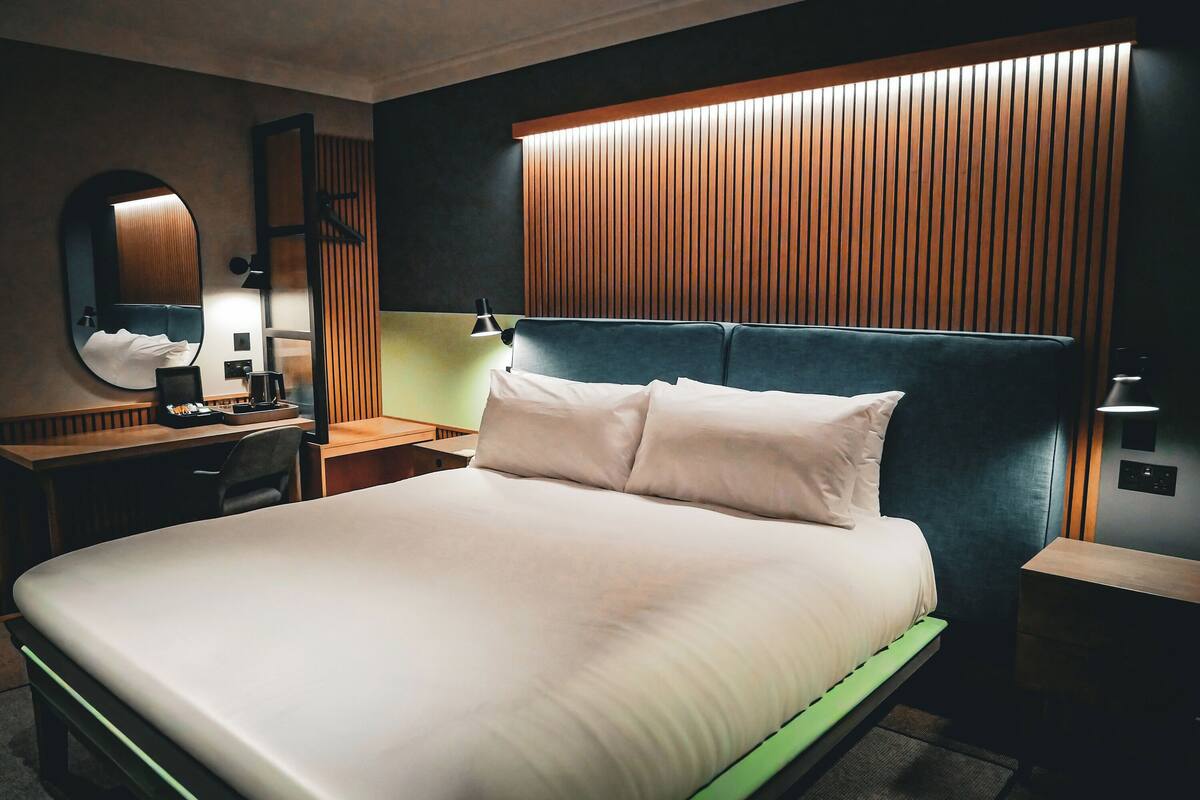 A modern hotel room with a large bed, a desk, a chair, a mirror, and a wooden panel wall.