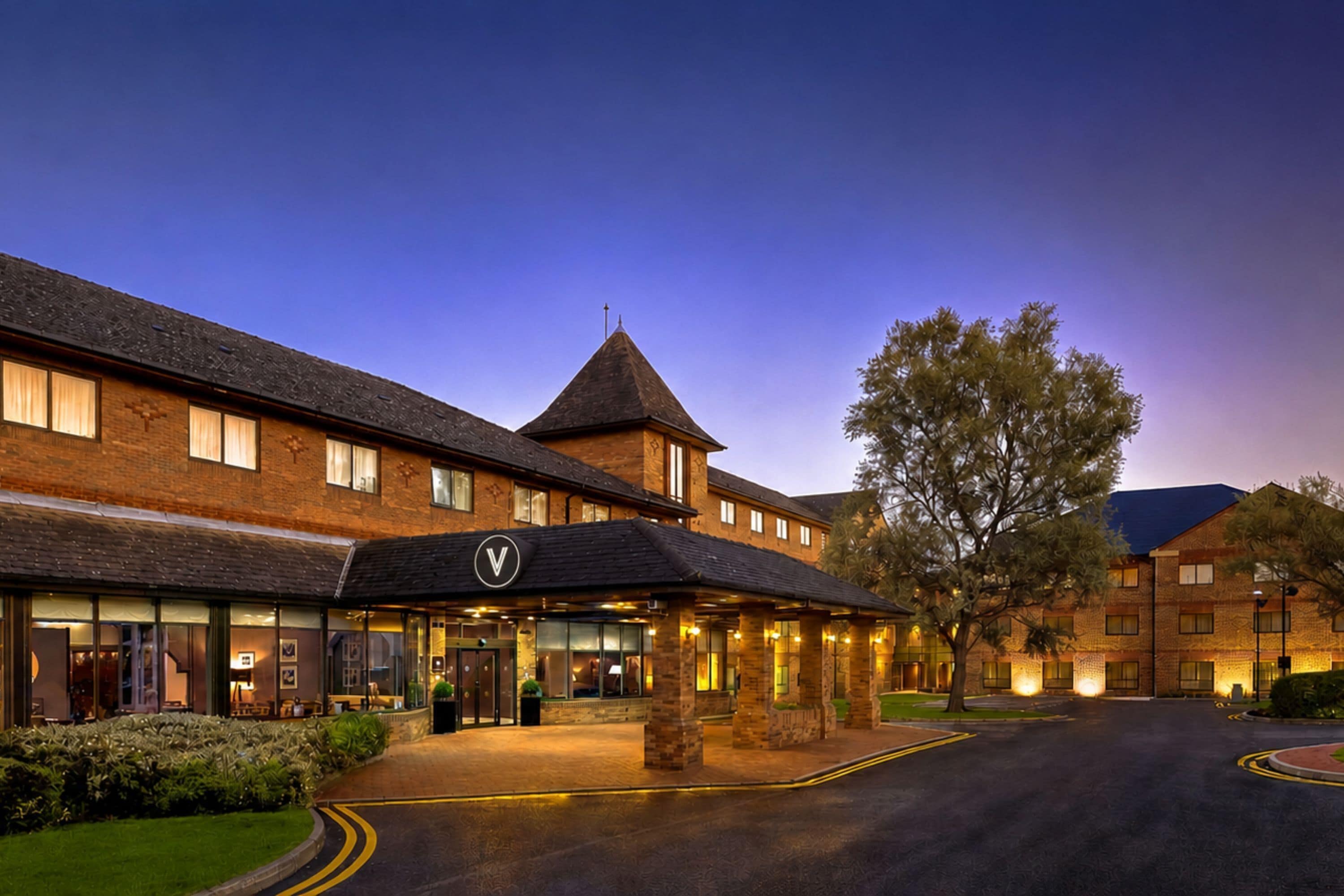 A hotel with a prominent entrance, a large tree in front, and a well-lit driveway.