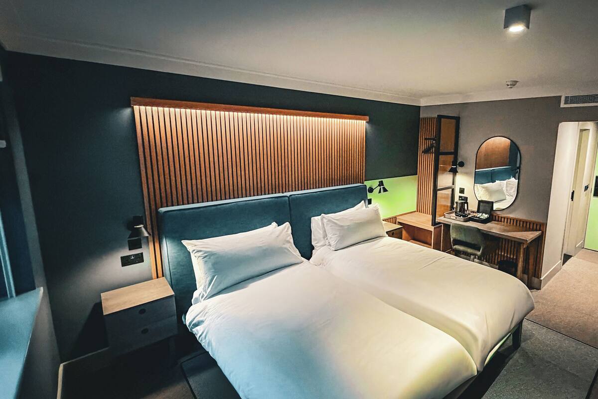 A modern hotel room with a large bed, bedside tables, a desk with a chair, a mirror, and a wooden headboard.