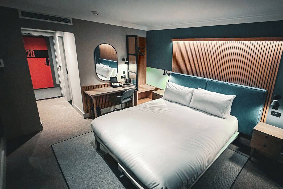 A modern hotel room with a large bed, a desk with a chair, a mirror, and a wardrobe.