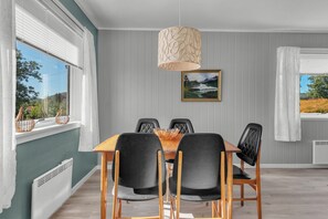 A dining area with a wooden table, black chairs, a hanging light fixture, and a framed landscape painting on the wall.