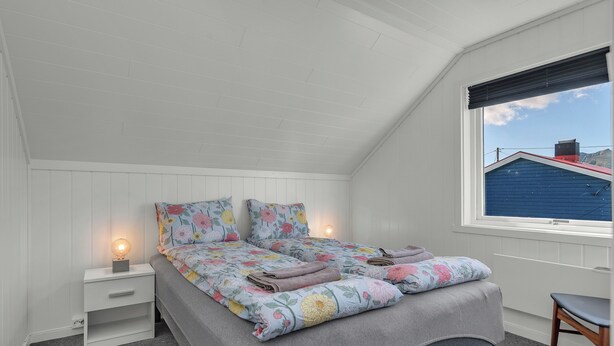 A bedroom with a sloped ceiling, a bed with floral bedding, a bedside table with a lamp, and a window with a view of a blue building.