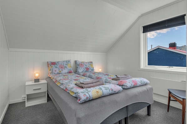 A bedroom with a sloped ceiling, a bed with floral bedding, a bedside table with a lamp, and a window with a view of a blue building.