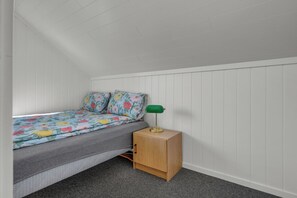 A small, single-bed room with a sloped ceiling, a wooden nightstand with a green lamp, and a floral-patterned bedspread. - Northern Norway Hidden Gem. Whales · Lights · Midnight Sun · Lofoten (Nordland)