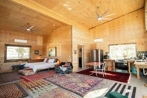 Interior de una cabaña de madera con cama, zona de estar, kitchenette y ventilador de techo.