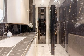 A modern bathroom with a large sink, a glass shower enclosure, and marble walls. - Luxury Apartment in Nice Plage (Nice)
