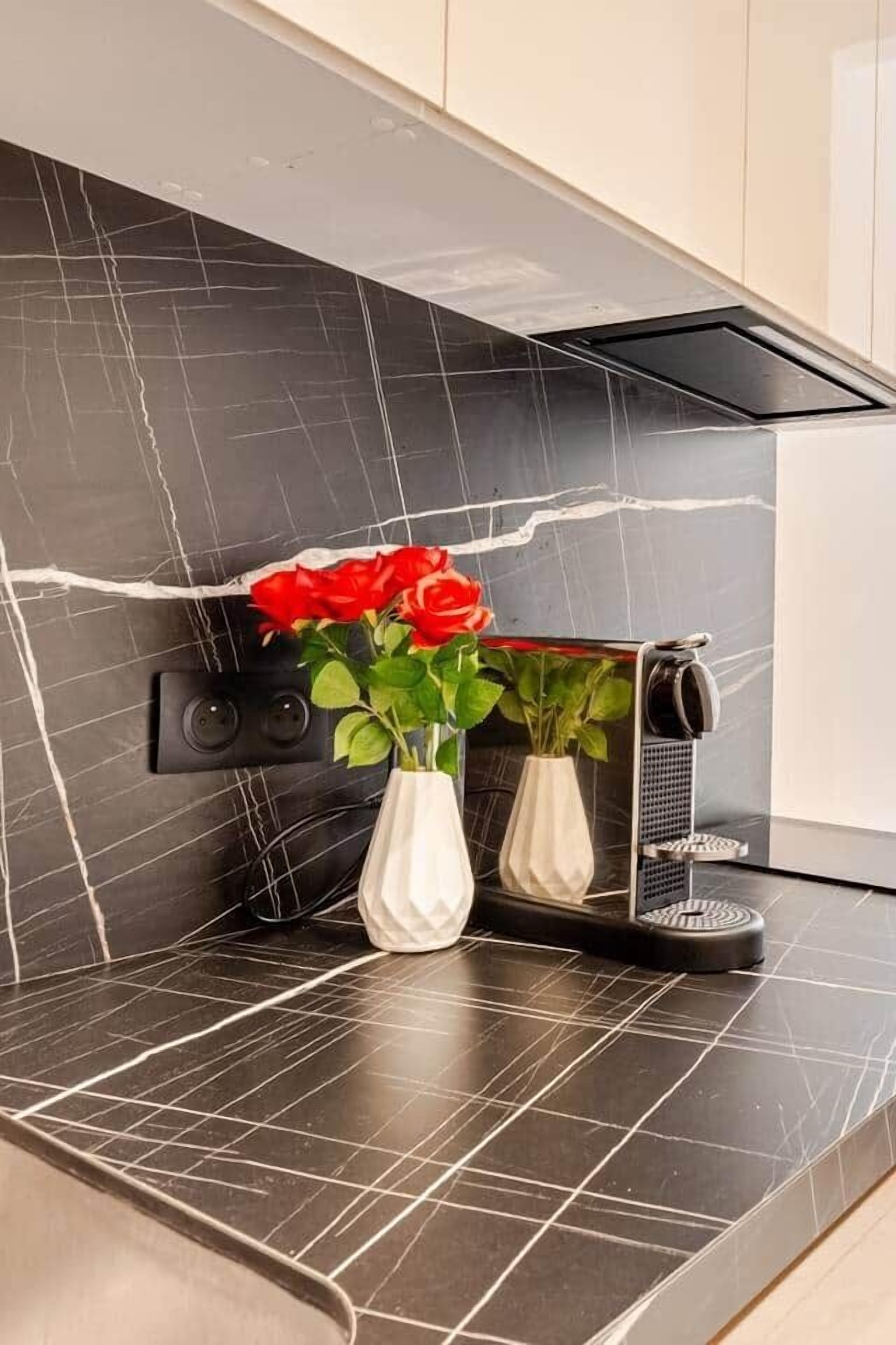 A kitchen counter with a black marble backsplash, a white vase with red roses, and a coffee machine.