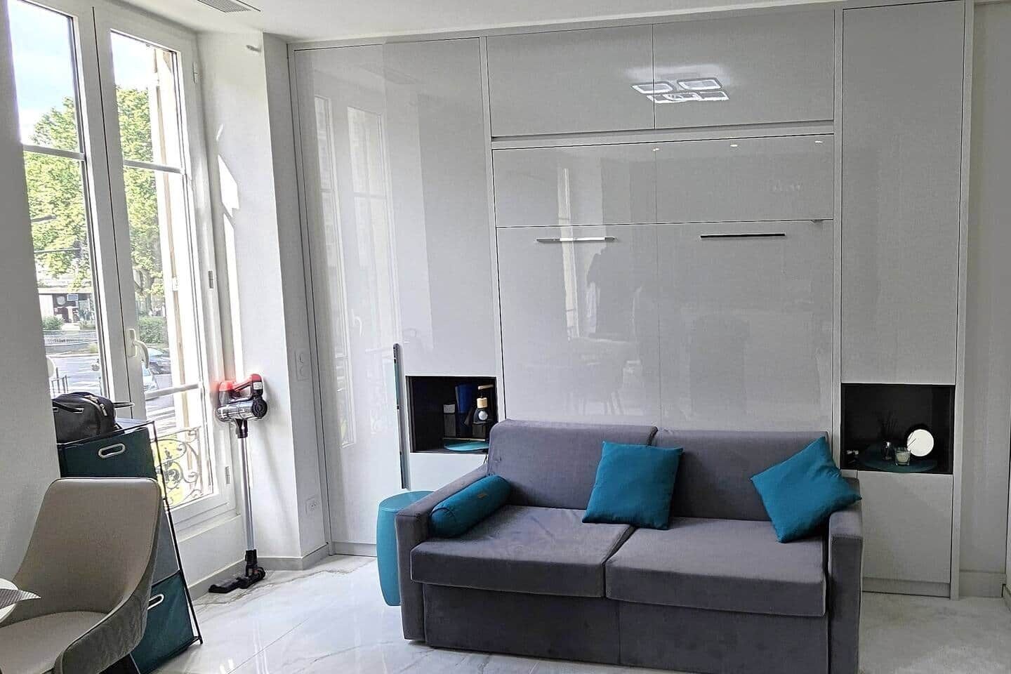 Un intérieur moderne avec un canapé gris, des coussins turquoise, une fenêtre offrant une vue et une porte dotée d’une poignée.