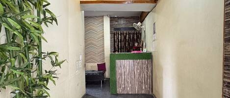 A narrow hallway with a reception desk, a bed, and a large potted plant.