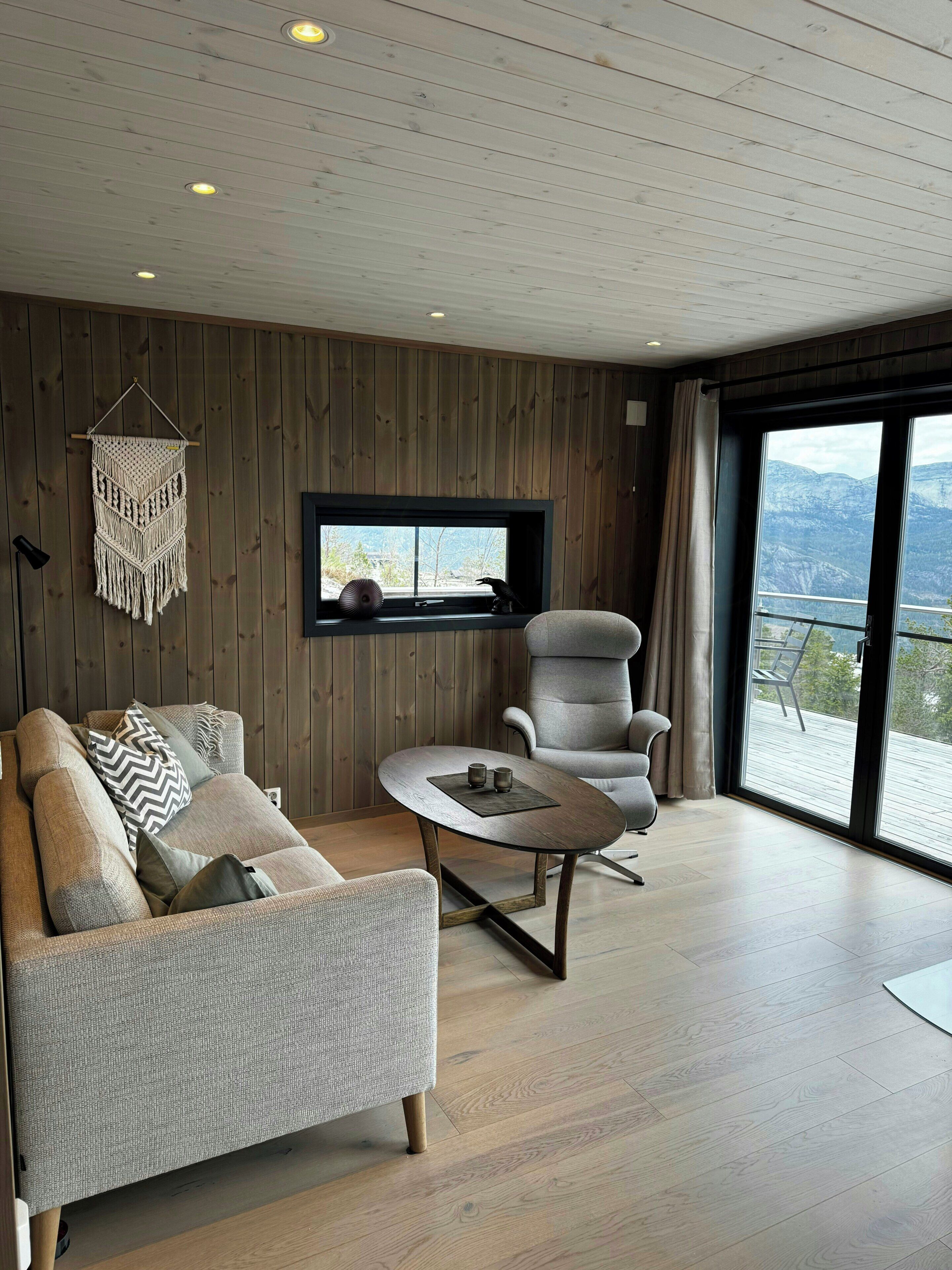 Ein modernes Wohnzimmer mit Sofa, Sessel und Couchtisch. Ein Wandbehang aus Makramee ist sichtbar, und es gibt ein Fenster mit Blick ins Freie.