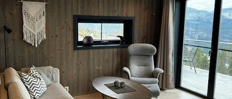 A modern living room with a sofa, armchair, and coffee table. A macrame wall hanging is visible, and there's a window with a view of the outdoors.