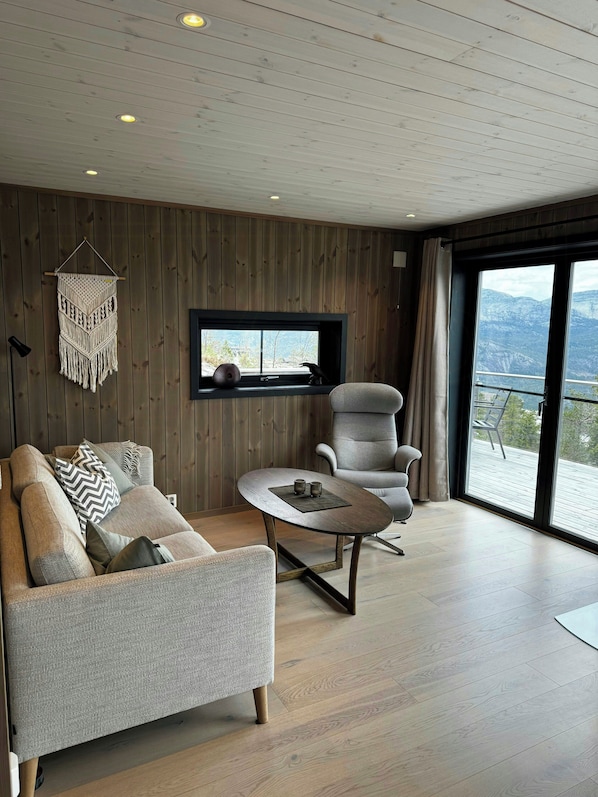 A modern living room with a sofa, armchair, and coffee table. A macrame wall hanging is visible, and there's a window with a view of the outdoors. - Modern cabin with panoramic views in Vrådal (Vrådal)