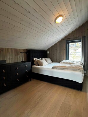 Ein Schlafzimmer mit schrägen Wänden, einem Bett mit schwarzem Kopfteil, einem Nachttisch und einer Kommode mit Schubladen.