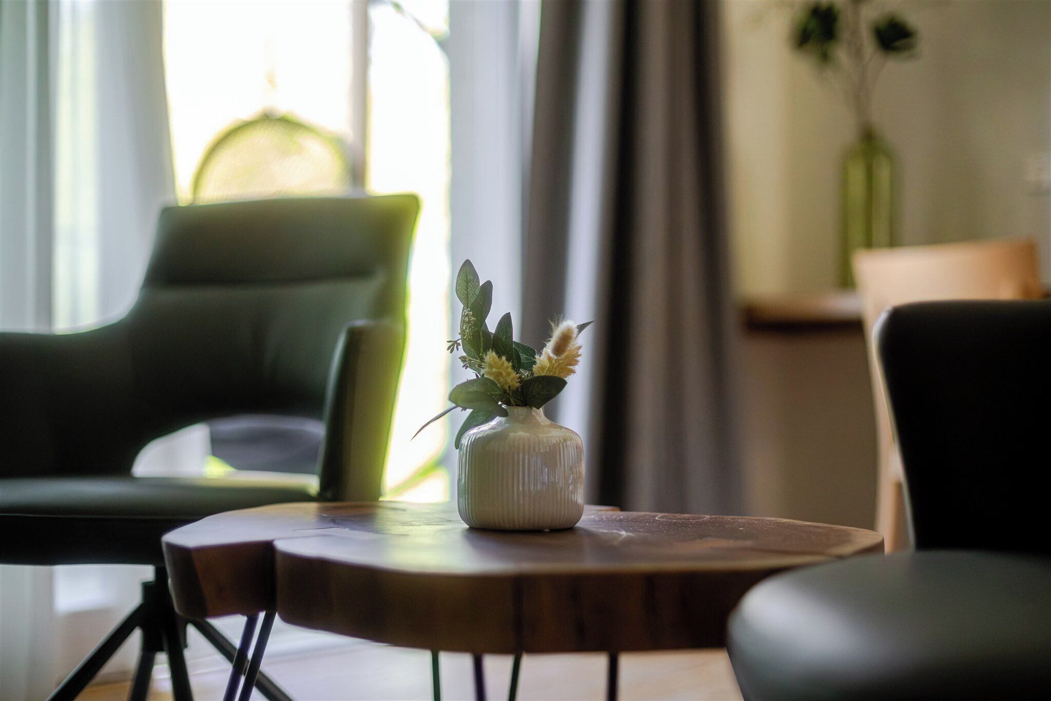 Una habitación con una mesa redonda de madera, un sillón verde y un jarrón blanco con una planta.