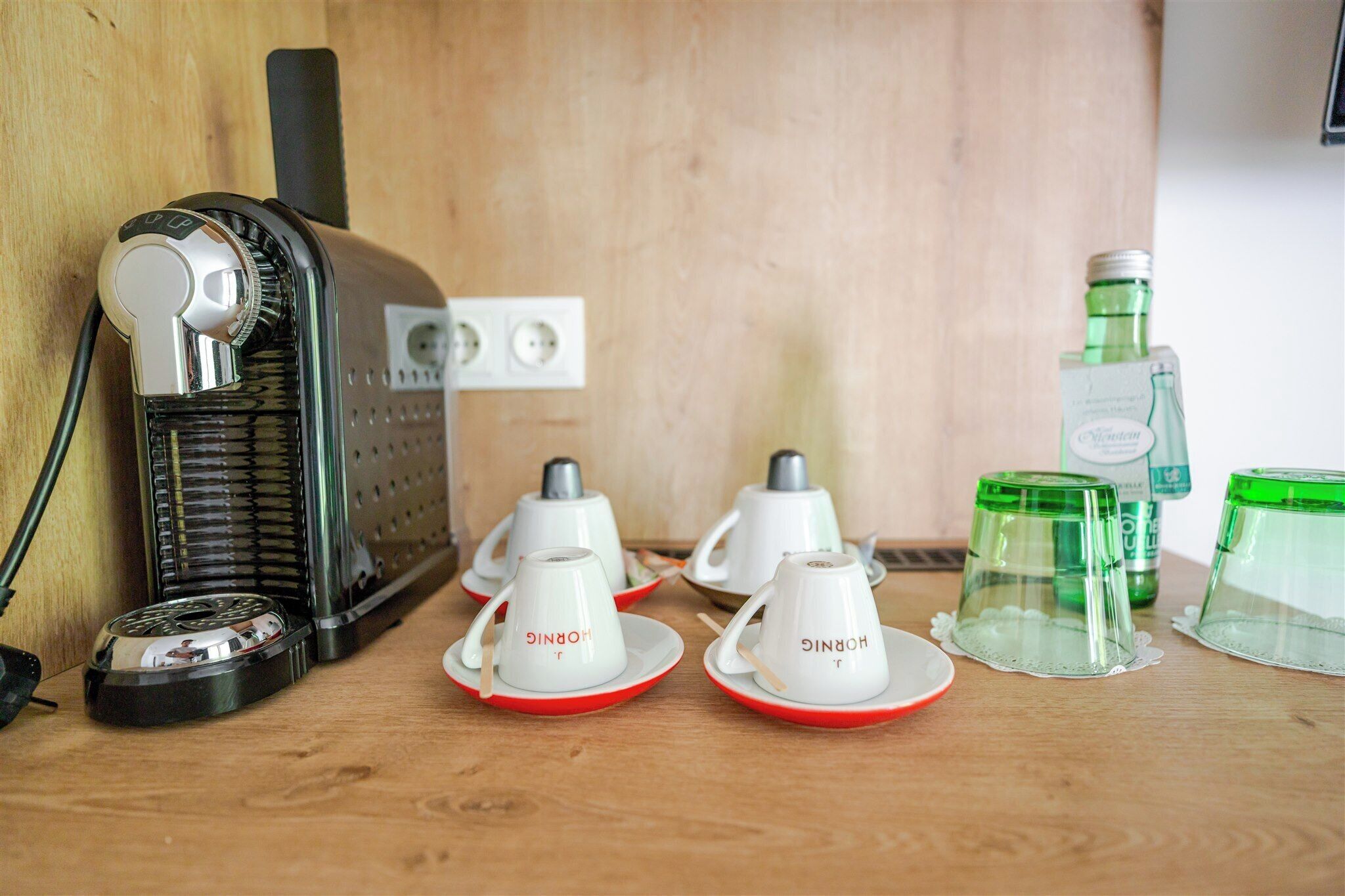 Una zona de cocina con cafetera, dos tazas blancas con detalles rojos y botellas de vidrio verde.
