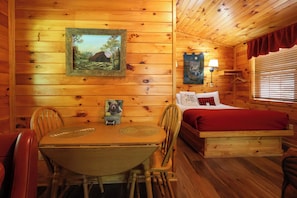 A wooden cabin interior with a dining table, chairs, a bed with a red blanket, a wall-mounted lamp, and a framed picture of a landscape. - Hot tub, on the river, firepit, hiking trails! (Sevierville)
