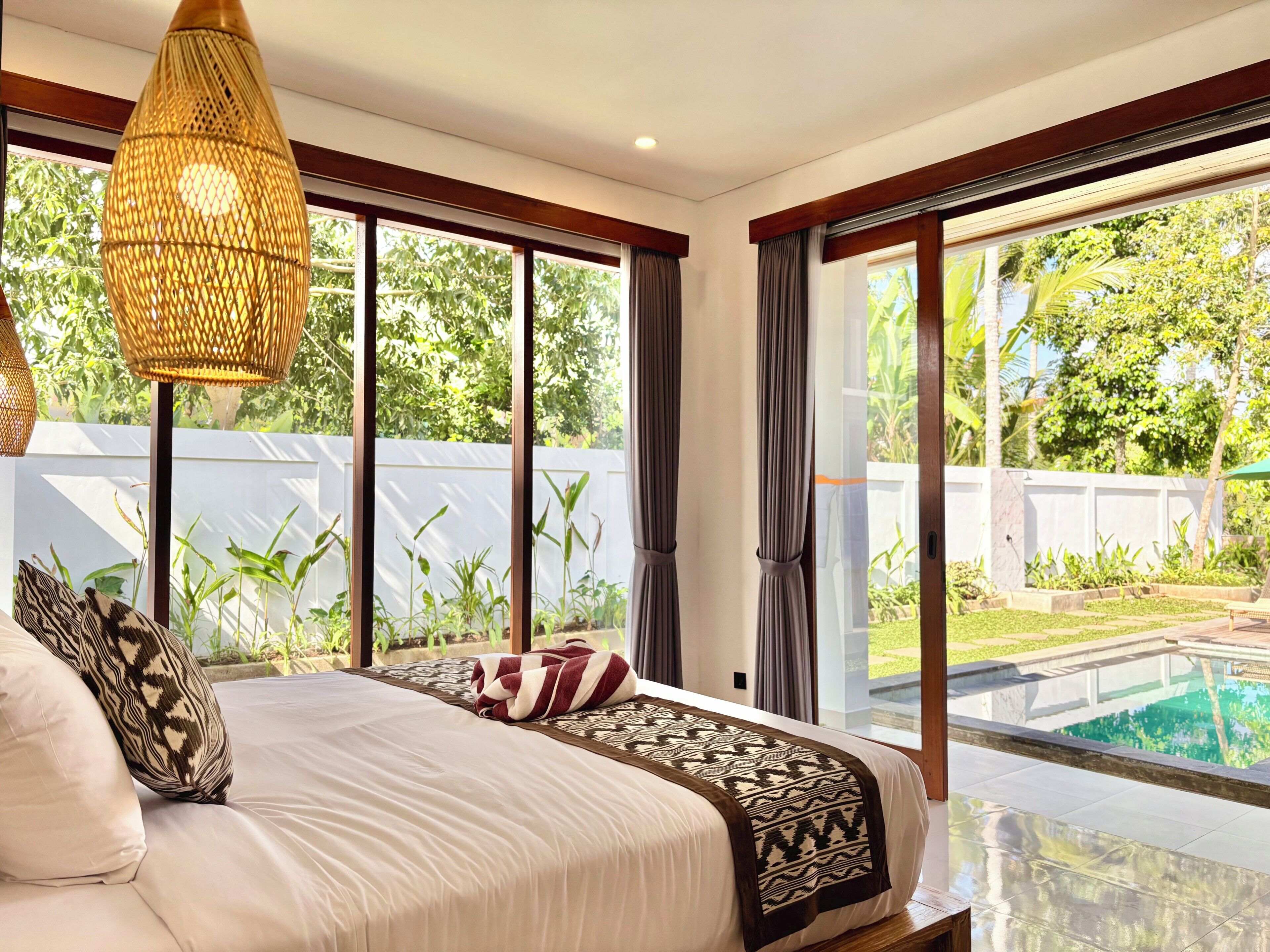 Ein Schlafzimmer mit einem großen Bett, Blick auf einen Swimmingpool und einem Balkon mit Pflanzen.