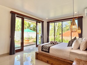 A bedroom with a large bed, a wooden headboard, and a view of a garden through large sliding doors. - 2-bedroom house in pleasant Ubud with cool AC for relaxing stay (Ubud)