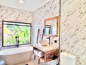 A bathroom with a freestanding bathtub, a wooden vanity with a bowl sink, a large mirror, and a window with a view of greenery. - 2-bedroom house in pleasant Ubud with cool AC for relaxing stay (Ubud)