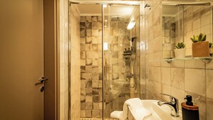 Un baño moderno con ducha de vidrio, un lavamanos blanco con dispensador de jabón negro y un espejo con repisa que sostiene una planta en maceta.
