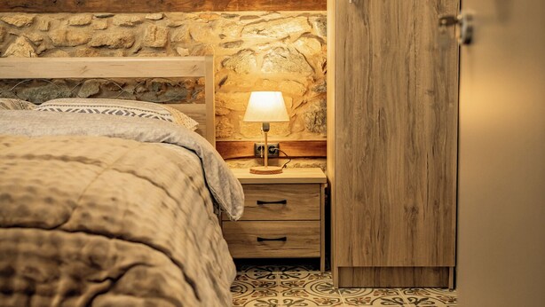 Un dormitorio con pared de piedra, cama de madera, mesita de noche con lámpara y alfombra roja estampada.